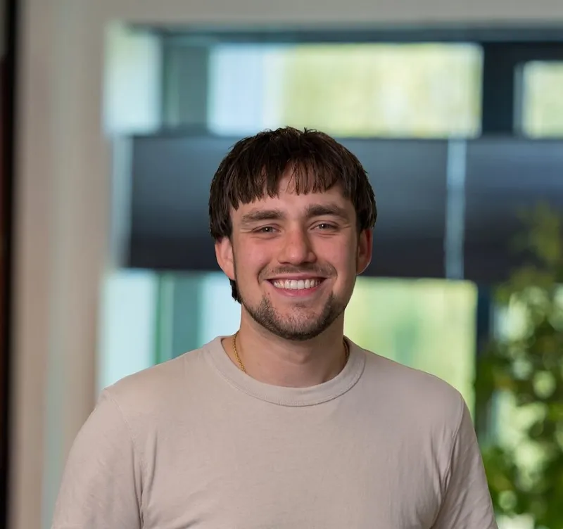 Portret van een jonge man met bruin haar, snor en sikje, gekleed in een beige T-shirt, glimlachend in een licht modern kantoor van Twyzer.