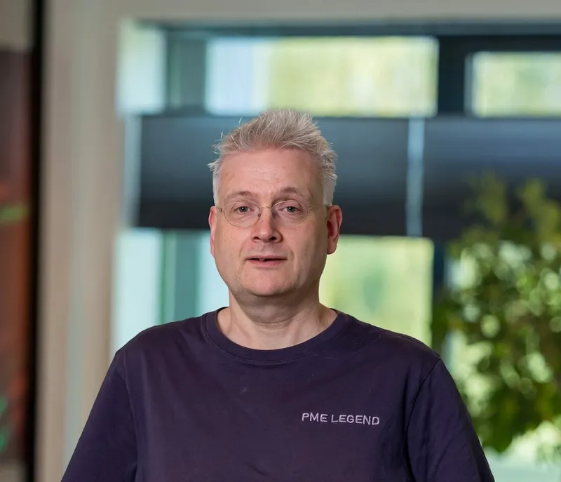 Portret van een man van middelbare leeftijd met kort grijs haar, bril en een donkerblauw T-shirt, staand neutraal kijkend op het Twyzer kantoor met ramen op de achtergrond.