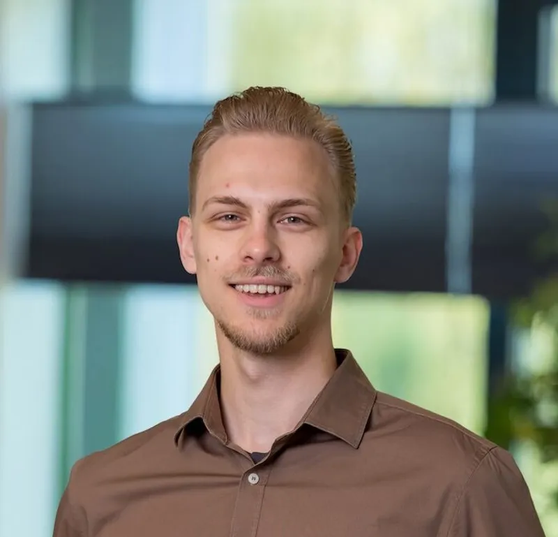 Portret van een jonge man met blondbruin haar en een sikje, gekleed in een bruin overhemd en jeans, zelfverzekerd glimlachend in een licht modern kantoor van Twyzer.