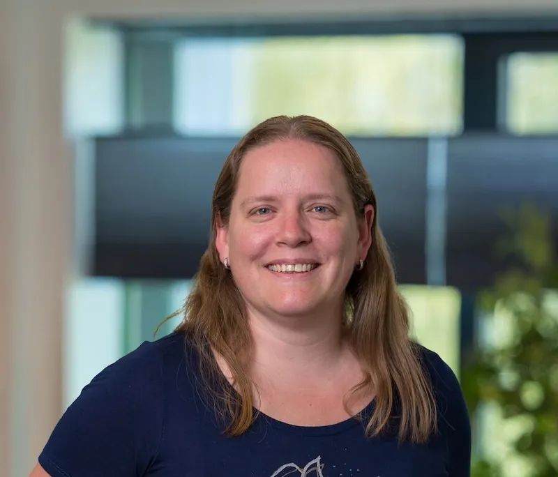 Portret van een vrouw van begin veertig met schouderlang blondbruin haar, gekleed in een donkerblauw T-shirt met bloemenprint en jeans, glimlachend op het Twyzer kantoor.