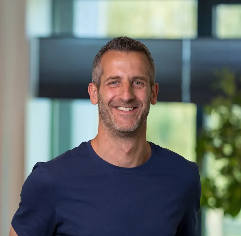 Portret van een man van begin vijftig met kort grijsbruin haar en baard, gekleed in een donkerblauw T-shirt, glimlachend met handen op de rug op het Twyzer kantoor.