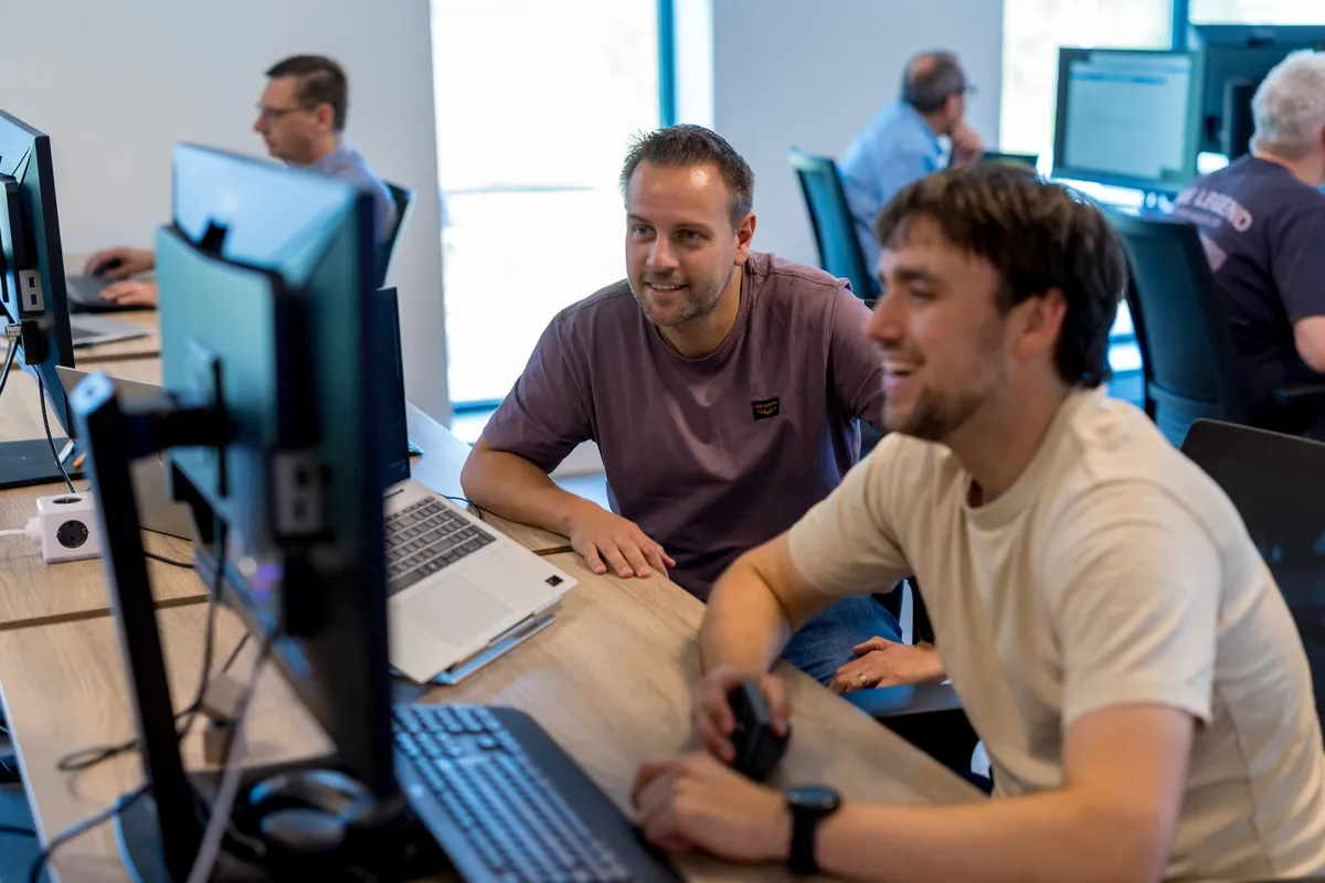Twee mannelijke medewerkers van Twyzer werken samen aan een computerscherm en lachen; een jongere man in een licht T-shirt en een oudere man in een paars T-shirt op een licht kantoor met collega's op de achtergrond.