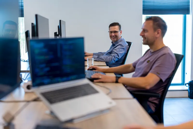 Hoe krijg je regie over data en technologie? Op het Twyzer kantoor werkt een man in een paars T-shirt aan een laptop met code op de voorgrond, terwijl een man van middelbare leeftijd met bril en blauw overhemd glimlachend over zijn schouder kijkt.