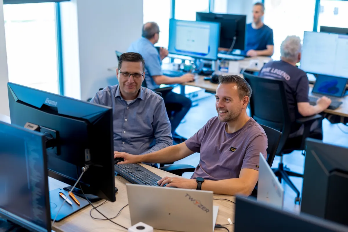 Teamwerk aan computers op kantoor Twee mannelijke medewerkers van Twyzer werken samen aan een bureau met meerdere schermen; een man met bril en blauw overhemd kijkt naar de camera terwijl een man in een paars T-shirt typt, met een laptop met Twyzer-logo op tafel.