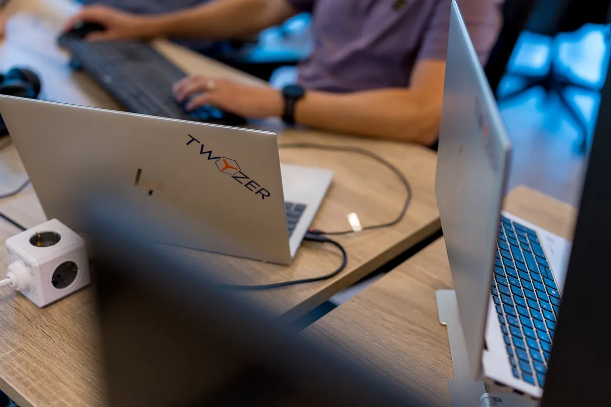 Close-up van een zilveren laptop met een Twyzer-sticker op een houten bureau, met op de achtergrond een medewerker in een paars poloshirt die aan een extern toetsenbord werkt.
