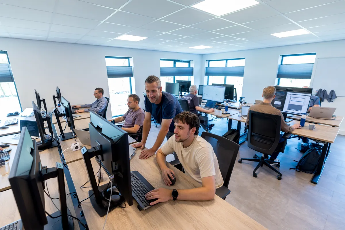 Overzicht van het lichte open kantoor van Twyzer met meerdere medewerkers achter grote schermen; op de voorgrond werken een jonge man in een crème T-shirt en een man met grijs haar in een blauw T-shirt samen aan een monitor.