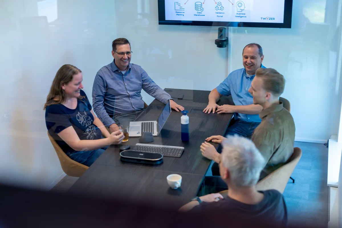 Vrolijk teamoverleg in vergaderzaal Vijf collega's zitten lachend rond een vergadertafel in een vergaderzaal met een Twyzer-presentatie op het scherm, gezien door een glazen wand.