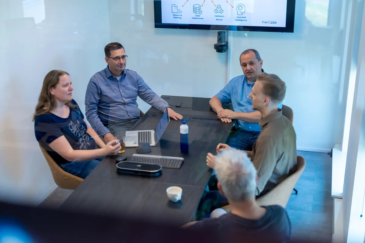 Teamvergadering achter glazen scheidingswand Vijf collega's zitten in een moderne vergaderzaal rond een tafel met laptop en conferentietelefoon, terwijl een technisch Twyzer-diagram op het scherm achter hen staat.
