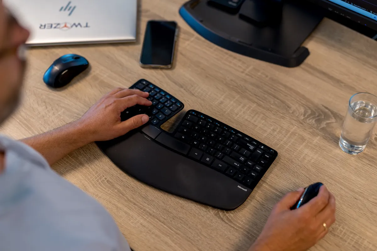 Over-de-schouder zicht op een medewerker die werkt met een Microsoft Sculpt ergonomisch toetsenbord en muis aan een houten bureau, met een laptop met Twyzer-logo op de achtergrond.