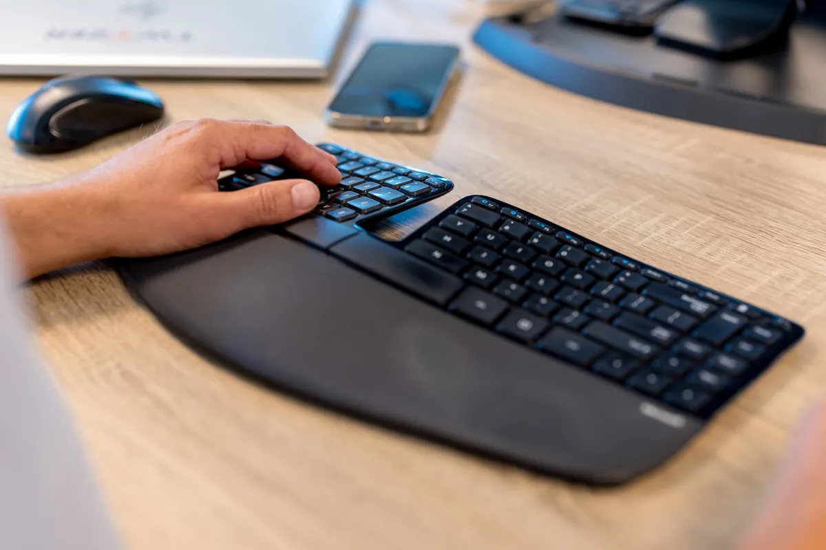 Handen op ergonomisch toetsenbord Close-up van een hand op een zwart ergonomisch split-toetsenbord op een houten bureau, met een muis, smartphone en monitor op de achtergrond.