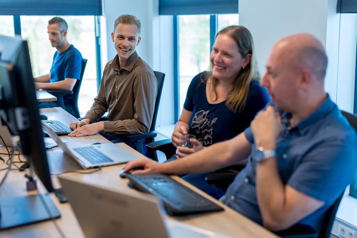 Vier medewerkers staan en zitten glimlachend bij hun bureaus in een licht kantoor en voeren een informeel gesprek met laptops en monitoren op de achtergrond.
