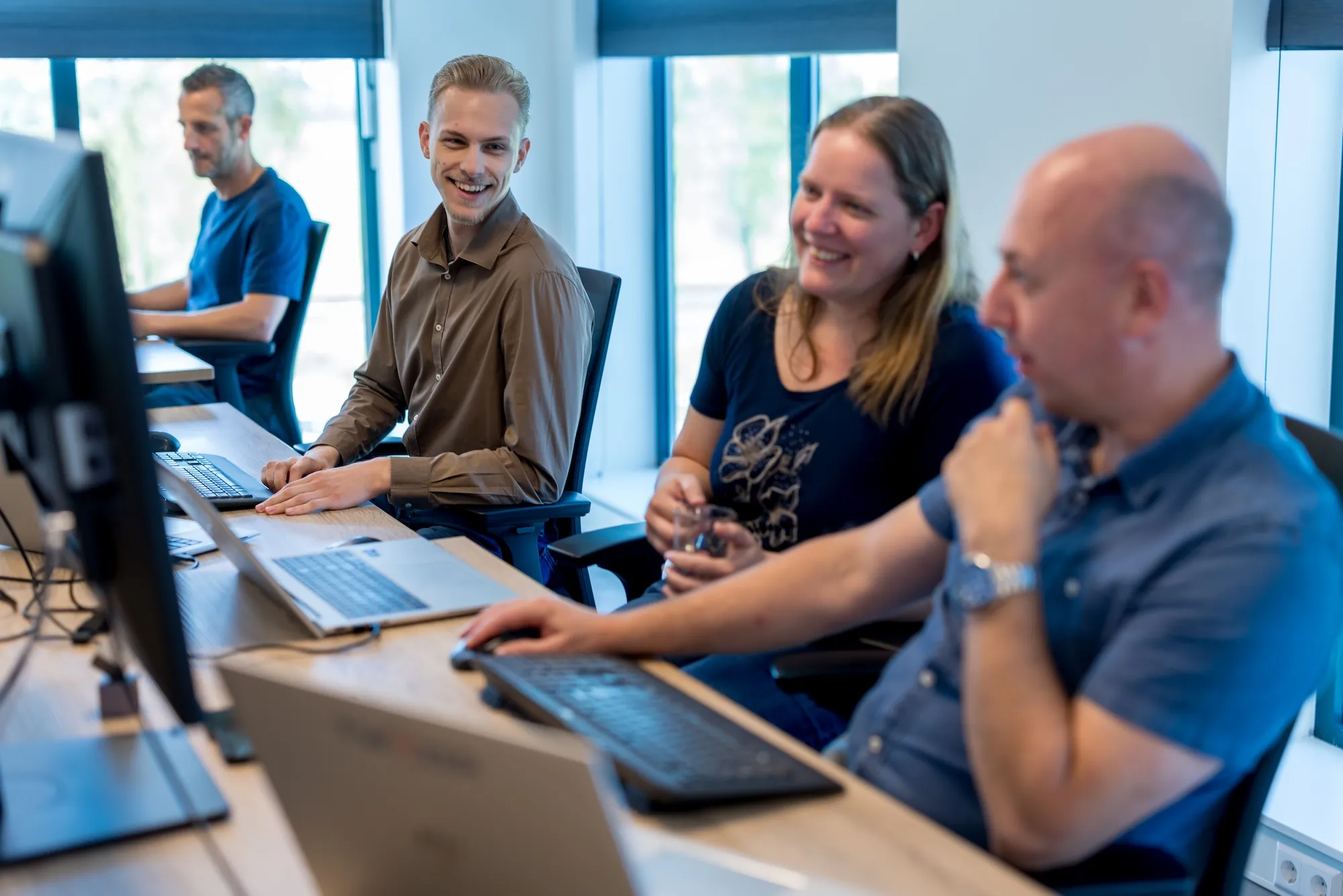 Collega’s in gesprek op kantoor Vier medewerkers staan en zitten glimlachend bij hun bureaus in een licht kantoor en voeren een informeel gesprek met laptops en monitoren op de achtergrond.