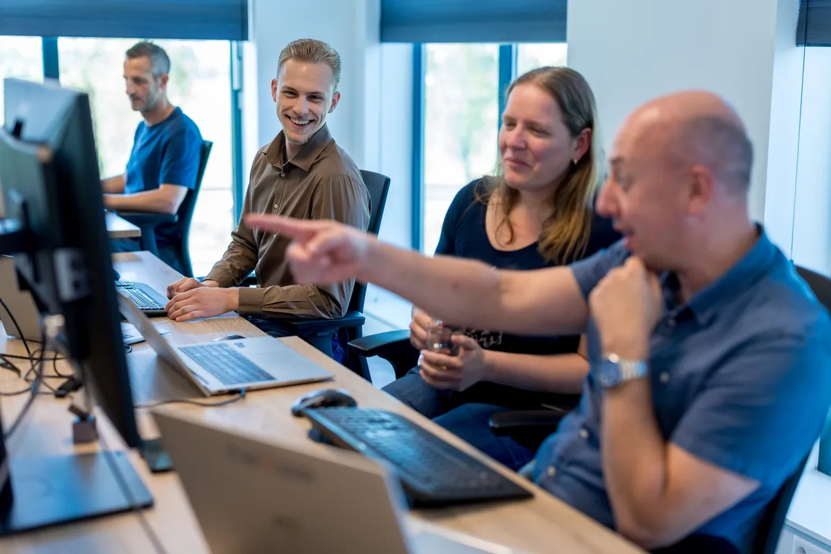 Uitleg aan collega bij computerscherm Een kale man wijst naar een computerscherm terwijl een vrouw met een glas en een jonge man glimlachend meekijken aan een bureau in een licht kantoor.