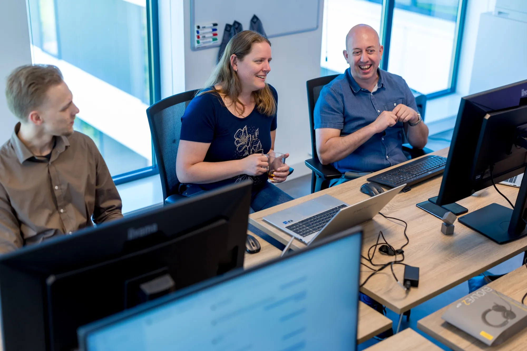 Lachende collega’s aan het bureau Drie collega's lachen en praten met elkaar aan een bureau met meerdere monitoren, laptops en een koptelefoon in een modern kantoor.