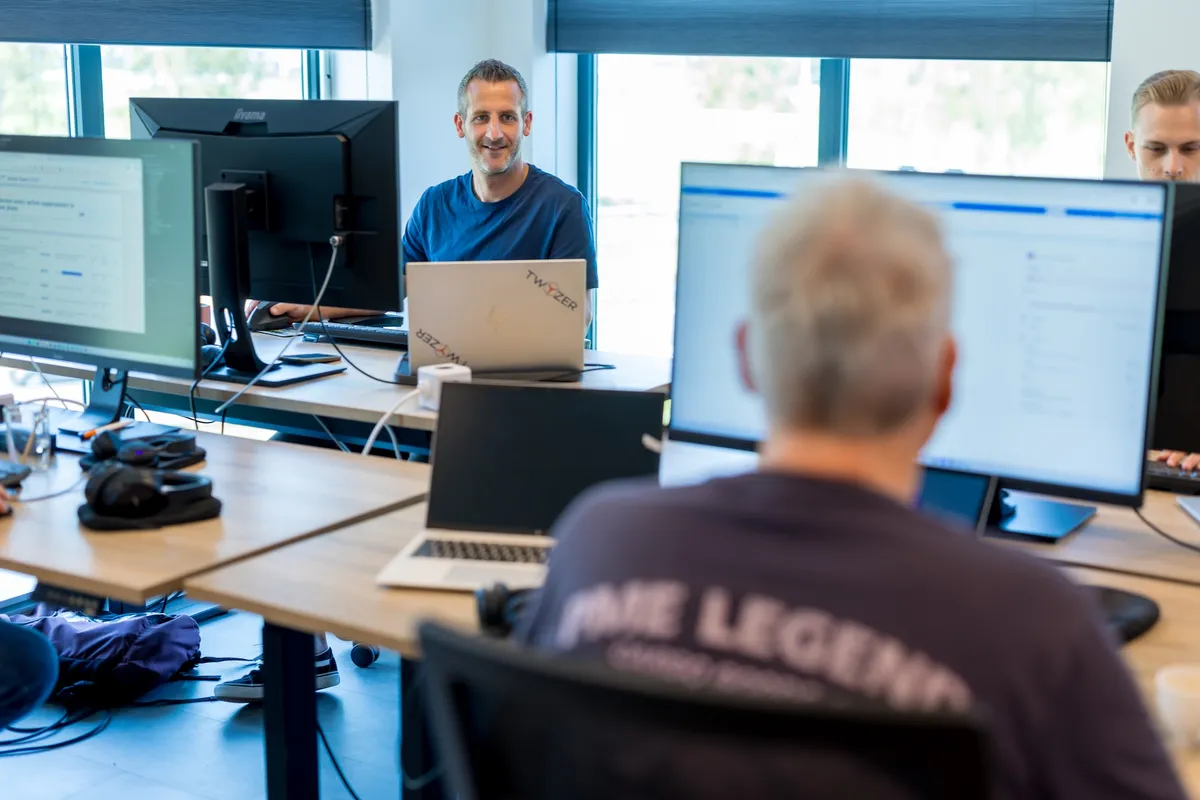 Een man in een blauw T-shirt zit glimlachend achter een laptop met Twyzer-stickers in een open kantoorruimte met grote ramen en blauwe jaloezieën.