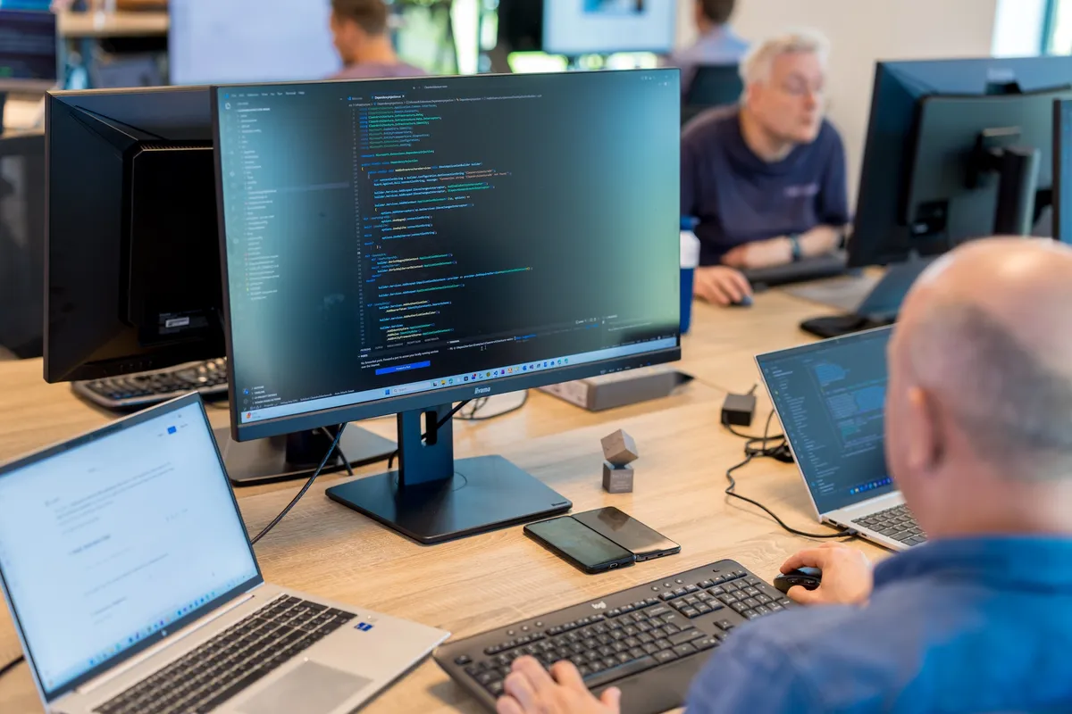 Meerdere schermen met broncode Een softwareontwikkelaar werkt aan een bureau met meerdere schermen waarop code en documentatie te zien zijn, terwijl collega's op de achtergrond aan het werk zijn.