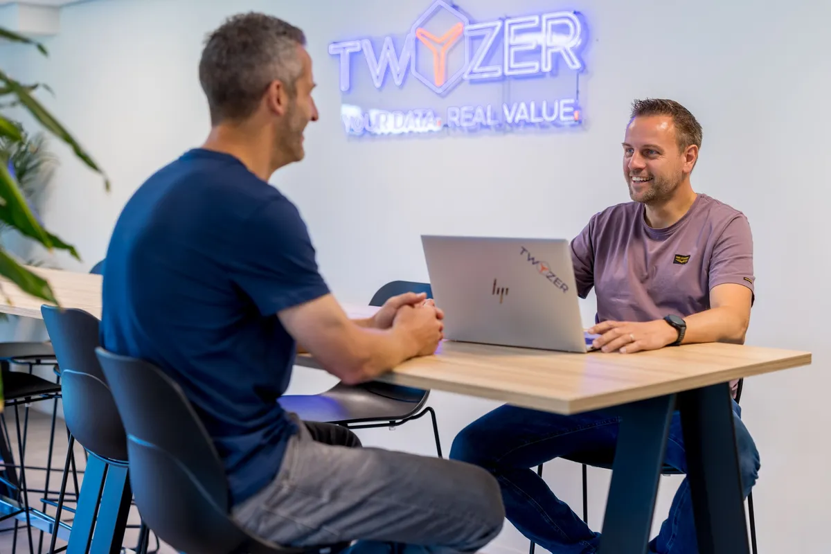 Samenwerking bij Twyzer Perry Lambrechts en een collega in gesprek aan een tafel in het Twyzer-kantoor met neon bedrijfslogo