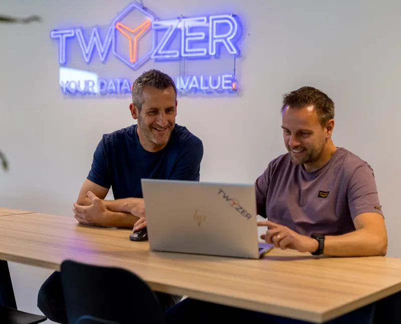 Don van Meel en Perry Lambrechts werken samen achter een laptop in het Twyzer-kantoor met neon logo op de achtergrond
